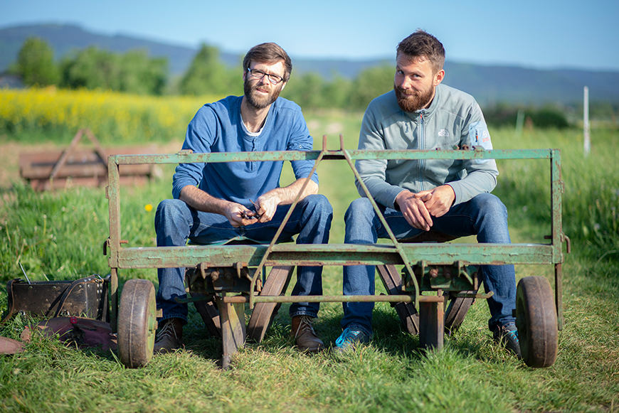 Die beiden Gründer Silas Müller und Christoph Graul von der "Kooperative" sitzen nebeneinander auf einem landwirtschaftlichen Gerät auf einem grünen Feld.
