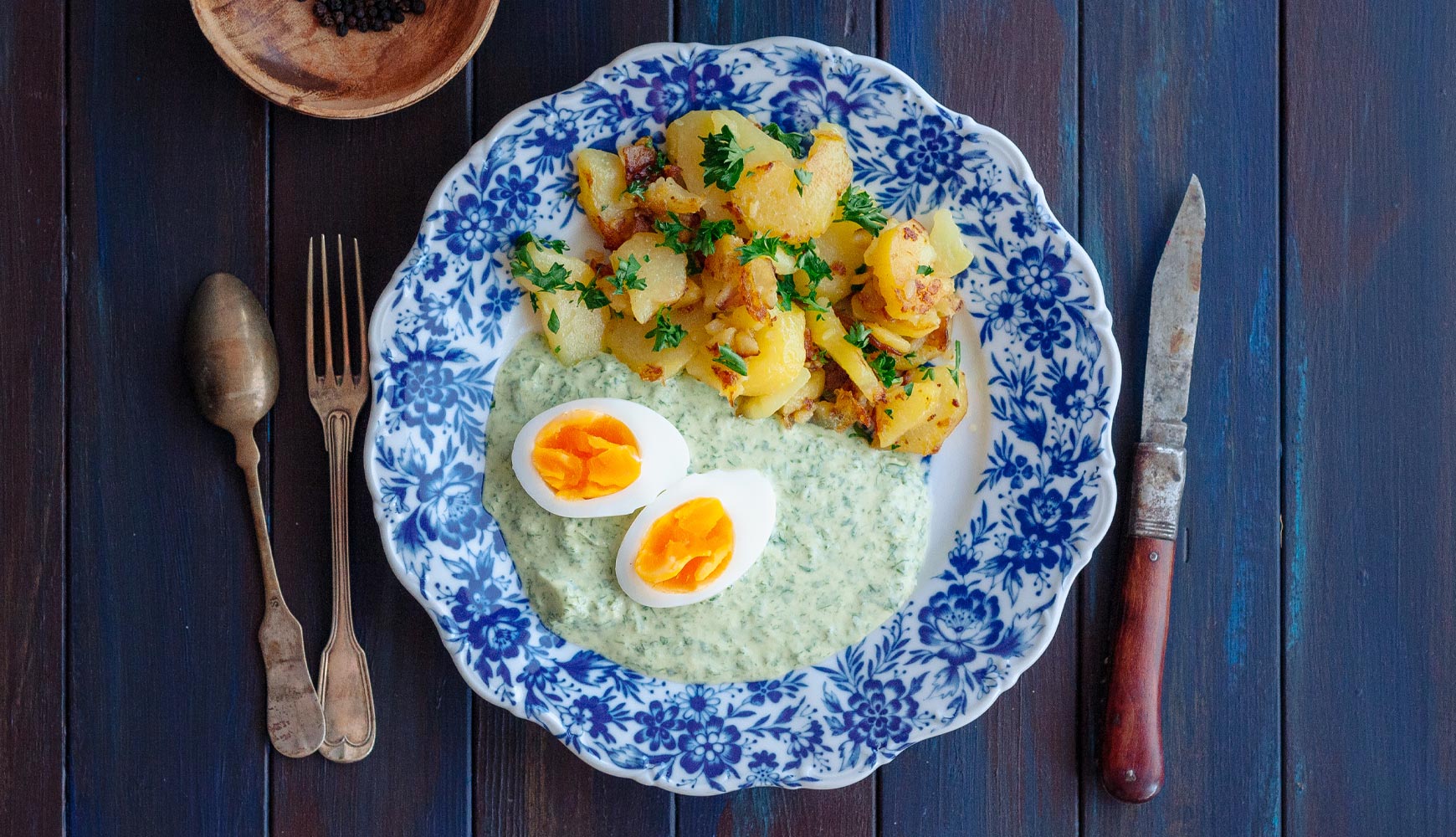 Ein Porzellanteller auf einem rustikalen, blauen Teller mit Bratkartoffeln und zwei halben Eiern in Frankfurter Grüner Spße
