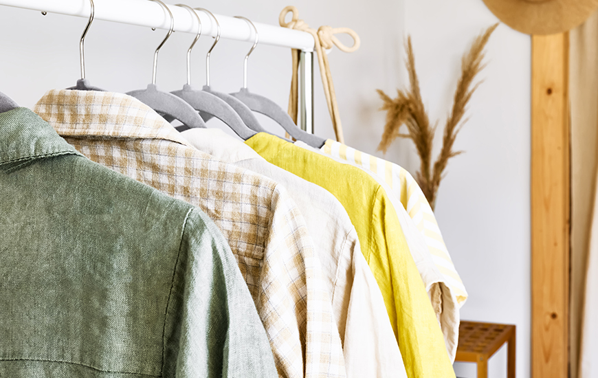 Eine Nahaufnahme einer Kleiderstange mit 5 Kleidungsstücken. Diese sind in gelb, hellbrauner und grüner Farbe.
