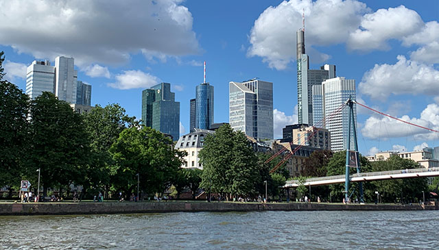 Auf dem Bild zu sehen ist die Skyline von Frankfurt. Davor sieht man das Mainufer mit spazierenden Menschen und vielen Bäumen. Am unteren Rand des Bildes ist der Main, rechts eine Brücke.