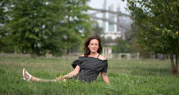 Feyza Morgül sitzt auf einer Wiese, dahinter die Frankfurter Skyline. Sie trägt ein schwarzes Kleid mit Muster und hat braunes gewelltes Haar.