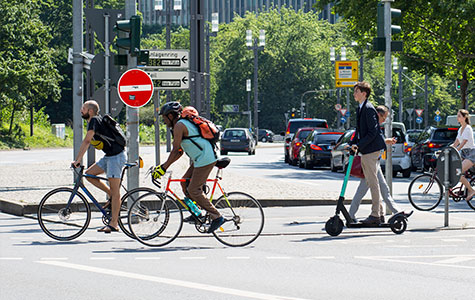 Eine Verkehrssituation einer Straßenkreuzung in Frankfurt. Man sieht drei Radfahrer, einen Fußgänger und eine Person auf einem E-Roller.