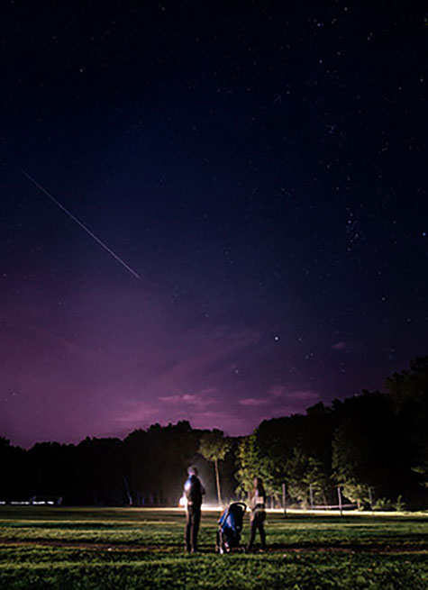 Ein Nachthimmel mit Sternen und einer Sternschnuppe. Auf der Wiese steht eine Familie mit Kinderwagen und schaut in den Himmel