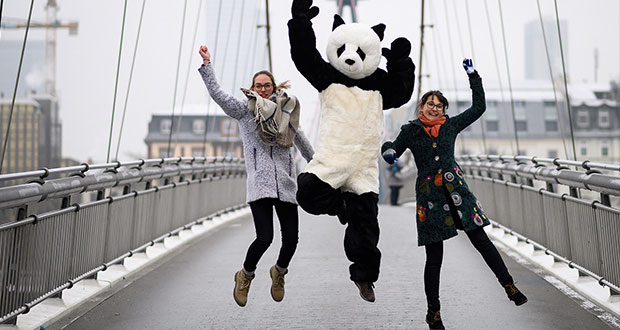 Zwei Frauen springen auf einer Brücke nach oben, und halten in der Mitte an ihren Händen einen Mensch in einem Panda Kostüm