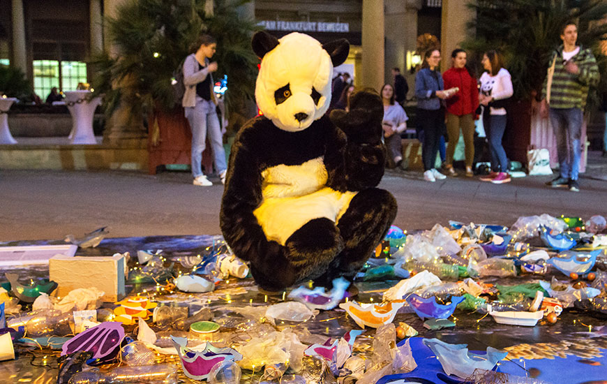 Ein Mensch in einem Panda Kostüm sitzt auf einem Haufen Plastikmüll und winkt in die Kamera