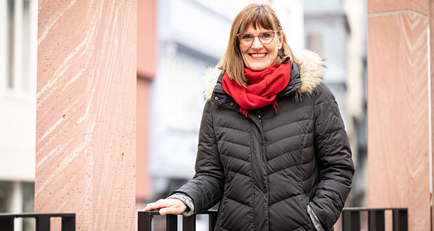 Bild von Karin Gerhardt: Sie steht an einem Geländer gelehnt mit schwarzer Winterjacke und roten Schal. Frau Gerhardt trägt eine Brille, hat braune schulterlange Haare und lächelt in die Kamera