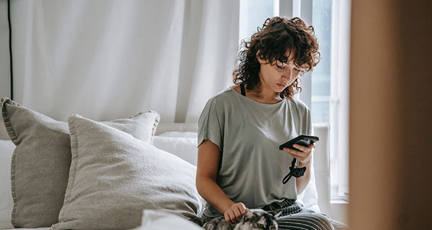 Eine junge Frau mit lockigem Haar seitzt auf ihrem Bett und schaut in ihr Smartphone