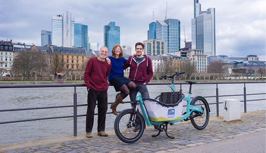 Manfred Fußnecker, Patricia Immler und Yannik Alff mit Lastenrad am Main. Im Hintergrund die Frankfurter Skyline.