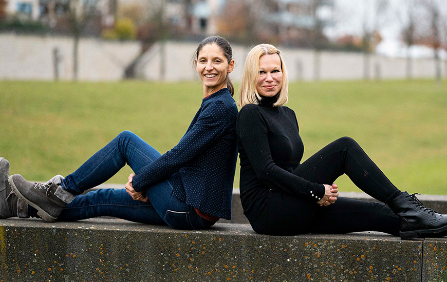 Die beiden Mitgründerinnen Ina Mirel und Anne Rave sitzen Rücken an Rücken auf einer Mauer und lächeln in die Kamera