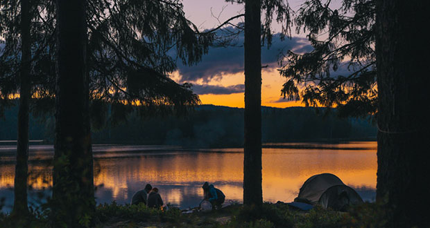 Ein Sonnenuntergang an einem See. Davor 3 Menschen am Lagerfeuer. Neben ihnen ein Zelt.