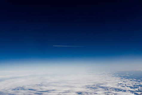 Ein Bild aus einem Flugzeug heraus. Man sieht Wolken, blauen Himmel und in weiter Ferne ein anderes Flugzeug