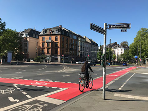 Eine große Straßenkreuzung an der Friedberger Landstraße in Frankfurt. Rot markiert sind die neuen Radspuren, darauf fährt eine Frau mit ihrem Fahrrad und kreuzt die Straße