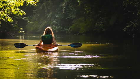 Eine Frau sitzt in einem Boot und paddelt über einen Fluss. Man sieht sie von hinten.