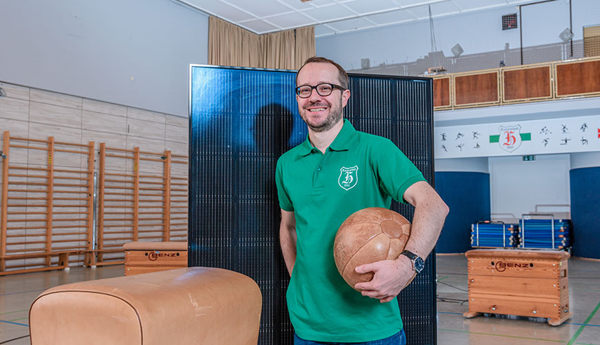 Portrait von Markus Schmid. Ein Mann mit grünem T Shirt, Brille und braunem Haar und Bart. Er lächelt in die Kamera und steht in einer Turnhalle mit einem Ball in der Hand. Dahinter ein Solarpannel.